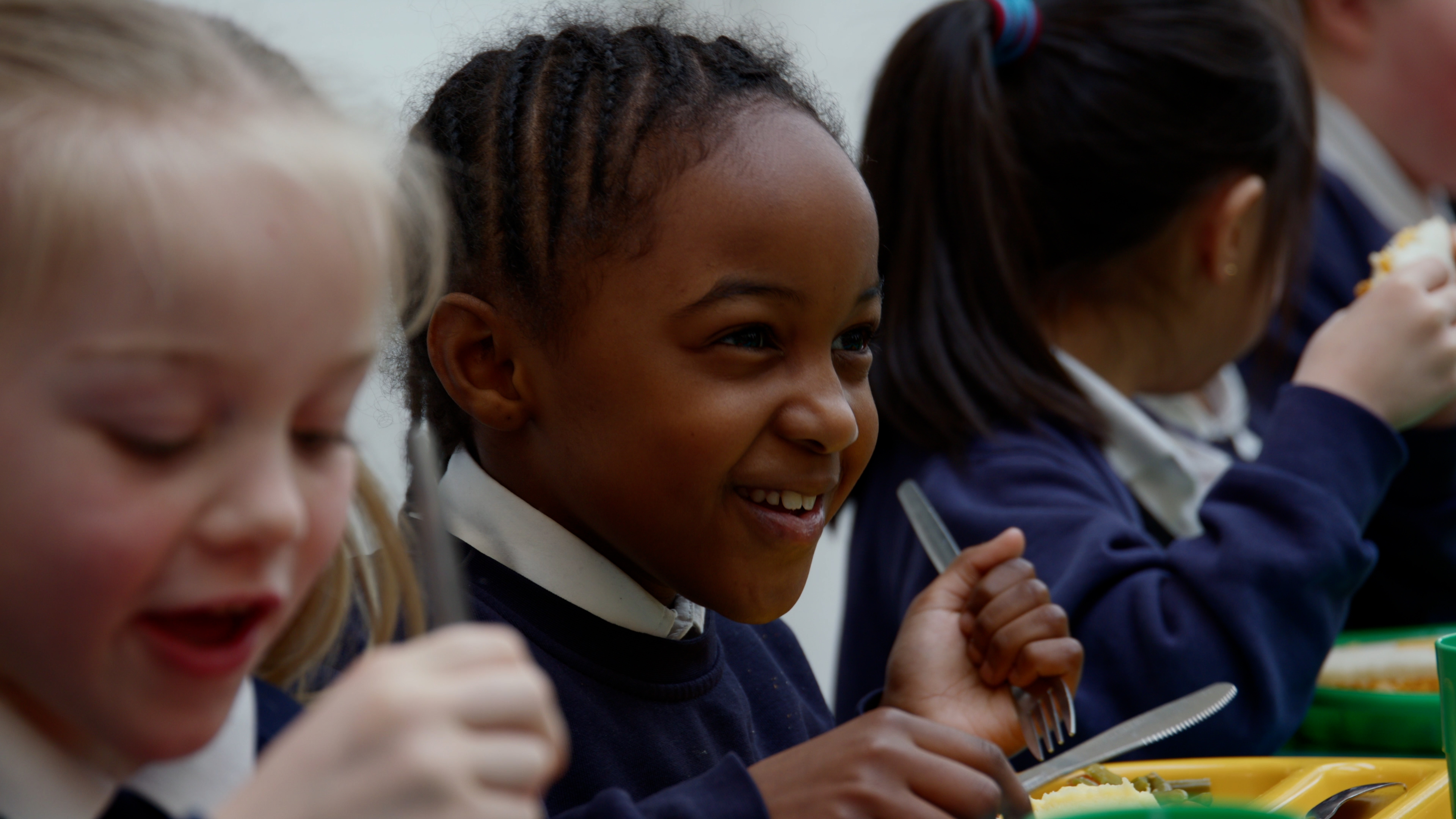 Three primary school aged children eating their school dinner. The middle child is laughing.